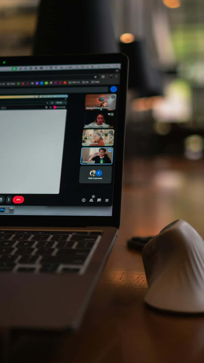 Na drevenom stole je položený notebook zobrazujúci videokonferenciu.